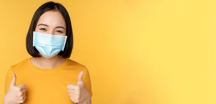 Cheerful korean woman in medical face mask, support people during pandemic, wear personal protective equipment from covid-19, showing thumbs up in approval, yellow background.