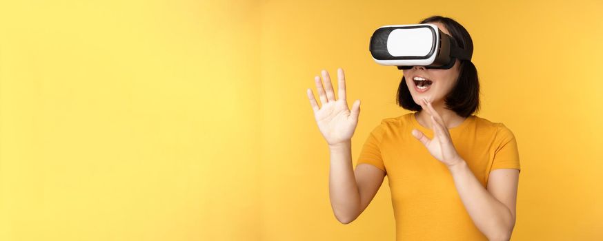 Girl in VR. Beautiful young asian woman, using virtual reality glasses and playing, chatting virtually, standing over yellow background.