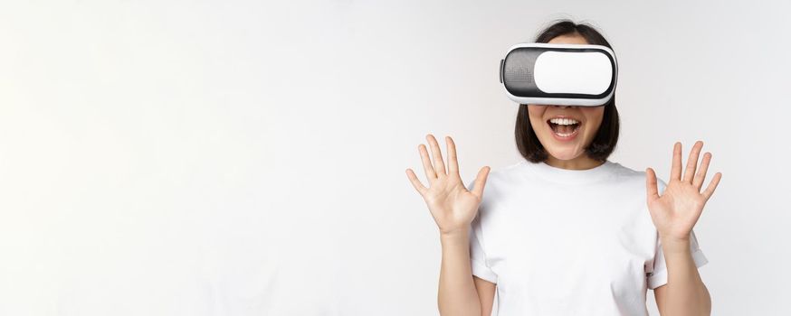 Happy asian woman using VR headset, waving raised hands and laughing, using virtual reality glasses, standing over white background.