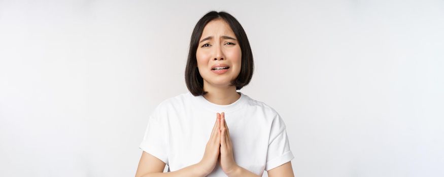 Desperate crying asian woman begging, asking for help, pleading and say please, standing in white t-shirt over white background.