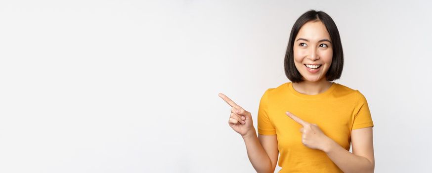 Portrait of smiling asian brunette girl in yellow tshirt, pointing fingers left, showing copy space, promo deal, demonstrating banner, standing over white background.