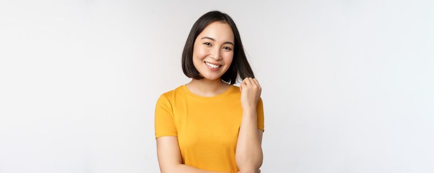 Beautiful romantic asian girl, smiling and playing with hair, looking happy at camera, standing in yellow t-shirt over white background.