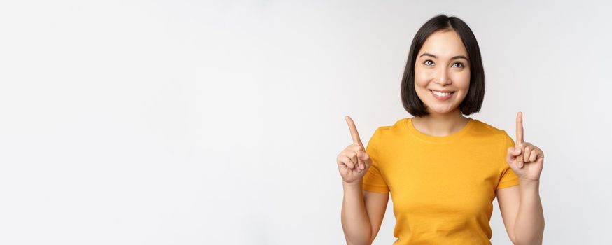 Happy beautiful asian girl showing advertisement, pointing fingers up, standing over white background.