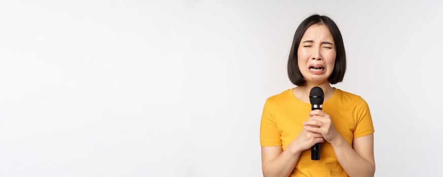 Crying asian girl singing heartbroken in microphone, holding mic and grimacing upset, standing over white background. Copy space