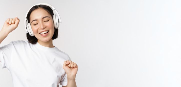 Dancing and singing asian woman, listening music in headphones, standing in earphones against white background. Copy space