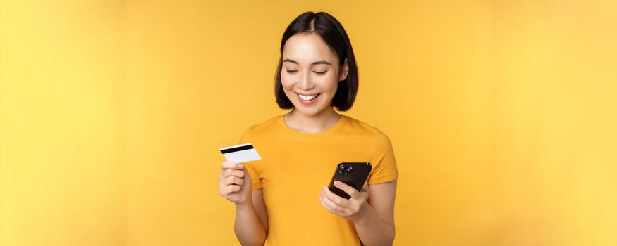 Online shopping. Smiling asian girl using credit card and mobile phone app, paying contactless, order on smartphone application, standing over yellow background.