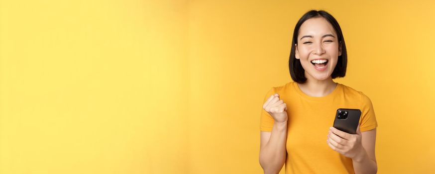 Joyful asian woman celebrating, holding mobile phone, winning, achieve goal on smartphone, standing over yellow background.