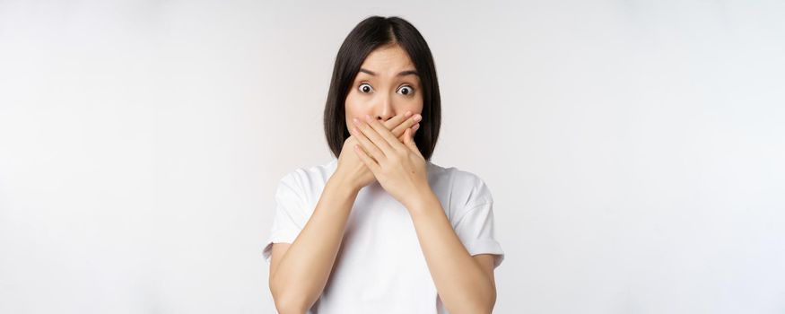 Image of shocked young asian woman shut mouth, looking startled, speechless, standing over white background. Copy space
