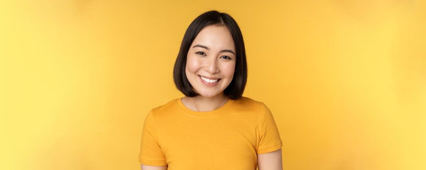 Close up portrait of beautiful asian woman smiling, looking cute and tender, standing against yellow background.