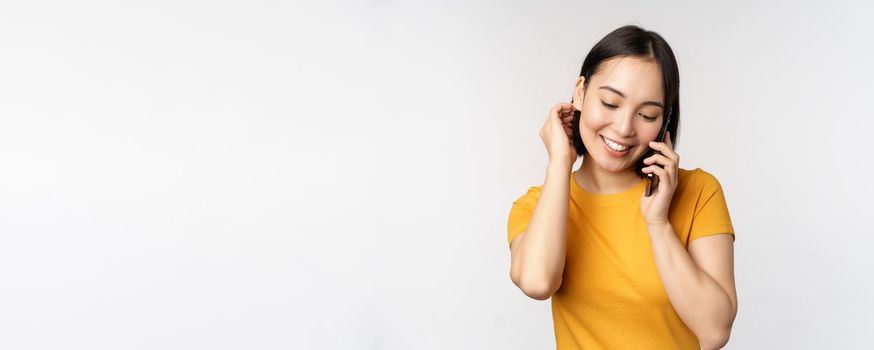 Romantic and cute asian girl answer phone call, talking on mobile, smiling coquettish, standing in yellow tshirt against white background.