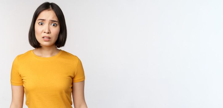 Portrait of worried korean girl, looking concerned at camera, standing in yellow tshirt over white background.