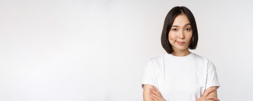 Close up of korean girl, looking skeptical, cross arms on chest and smirk, stare with disbelief at camera, standing over white background.