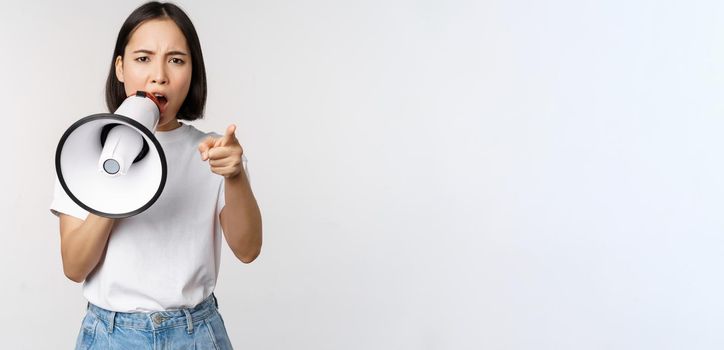 Angry asian woman with megaphone, scolding, accusing someone, protesting with speakerphone on protest, standing over white background.