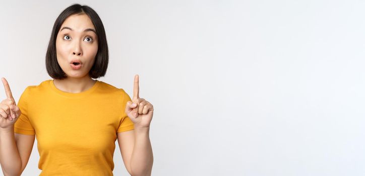 Happy beautiful asian girl showing advertisement, pointing fingers up, standing over white background.