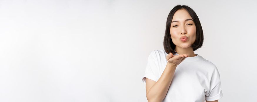 Lovely young asian woman, sending air kiss and looking coquettish at camera, standing in tshirt over white background.