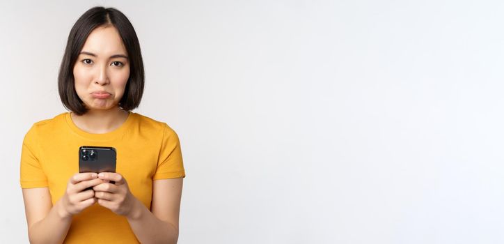 Sad asian woman holding smartphone, looking upset with regret, standing in yellow tshirt against white background.