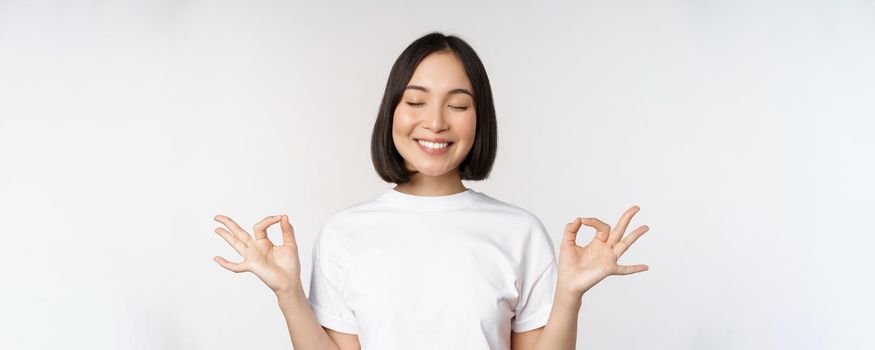 Portrait of young asian woman meditating, smiling pleased and practice yoga, close eyes and meditate, breathing calm, standing over white background.