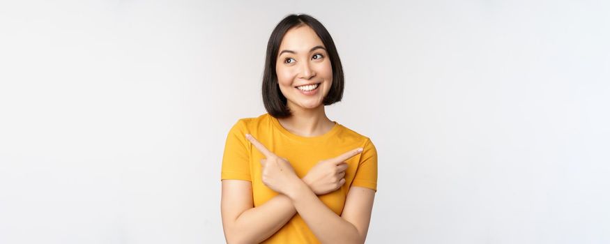 Cute asian girl pointing fingers sideways, showing left and right promo, two choices, variants of products, standing in yellow tshirt over white background.