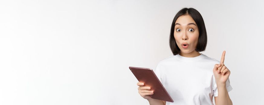 Enthusiastic asian woman with tablet, raising finger and looking amazed, pointing up, standing over white background.