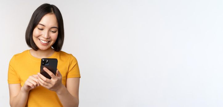 People and technology concept. Smiling asian girl using smartphone, texting on mobile phone, standing against white background.