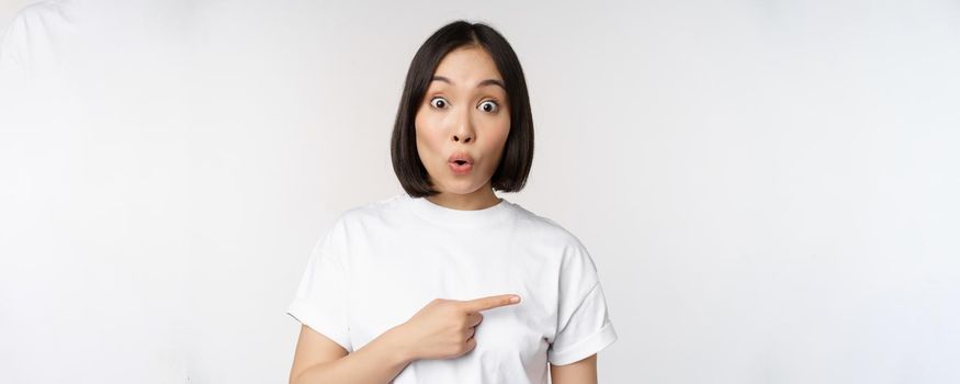 Portrait of surprised asian woman say wow, pointing right at amazing product offer, sale or logo, standing over white background.