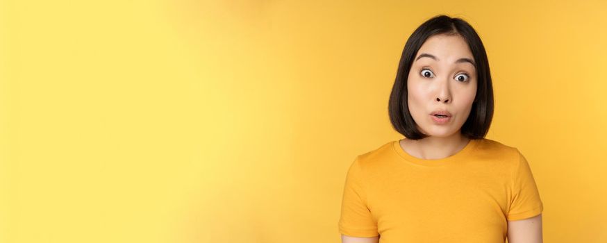 Close up portrait of asian girl showing surprised reaction, raising eyebrows amazed, reacting to big news, standing over yellow background.