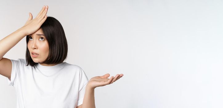 Annoyed korean girl facepalm, slap forehead and shrugging, confused by smth, standing over white background.