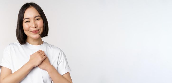 Portrait of carig young asian woman holding hands on heart, gazing with care and love, touched by something, feel flattered, standing over white background.