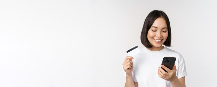 Online shopping. Happy asian woman using credit card and smartphone app, paying on website via mobile phone, white background.