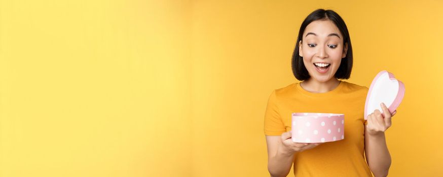 Portrait of excited asian woman, open gift box with surprised happy face, standing over yellow background.