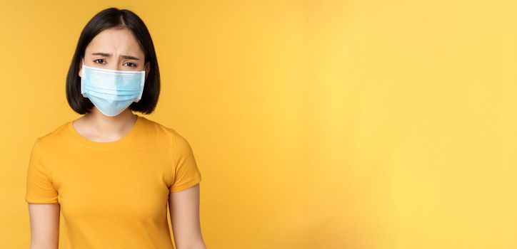 Image of sad and disappointed asian woman in medical mask, frowning and looking displeased, standing against yellow background.