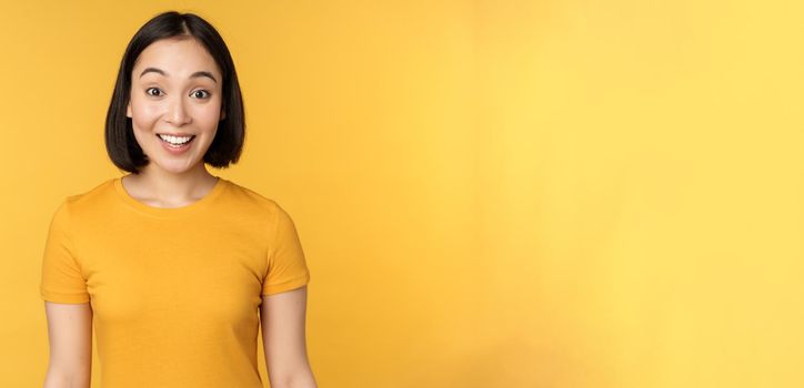 Image of asian girl looking surprised, reacting amazed, raising eyebrows impressed, standing over yellow background.