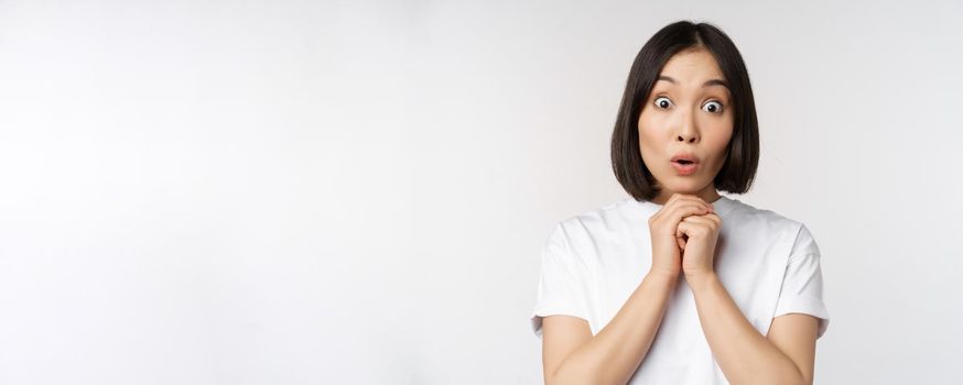 Close up portrait of asian brunette woman looking amazed, say wow, watching smth impressive, standing over white background.