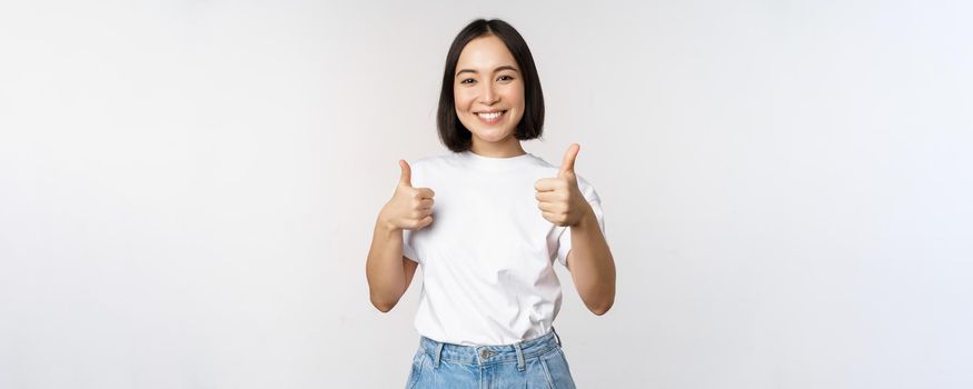 Happy beautiful korean girl, showing thumbs up in approval, smiling pleased, like smth, recommending, standing over white background.