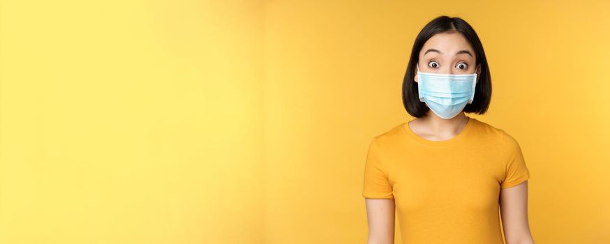 Portrait of korean girl in medical face mask looking surprised, amazed reaction to news, standing over yellow background.