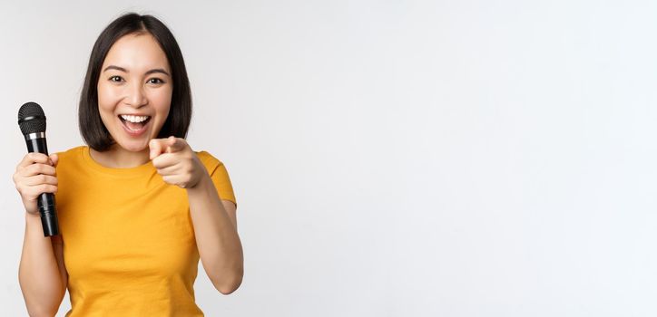 Enthusiastic asian girl with microphone, pointing finger at camera, suggesting you to sing, standing over white background.