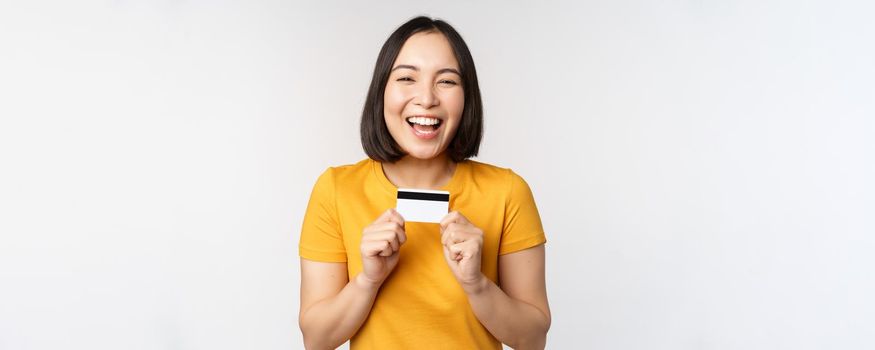 Portrait of beautiful korean girl holding credit card, recommending bank service, standing in yellow tshirt over white background.