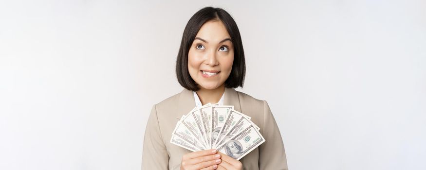 Image of asian corporate woman, happy businesswoman showing money, cash dollars and thinking, standing in suit over white background.