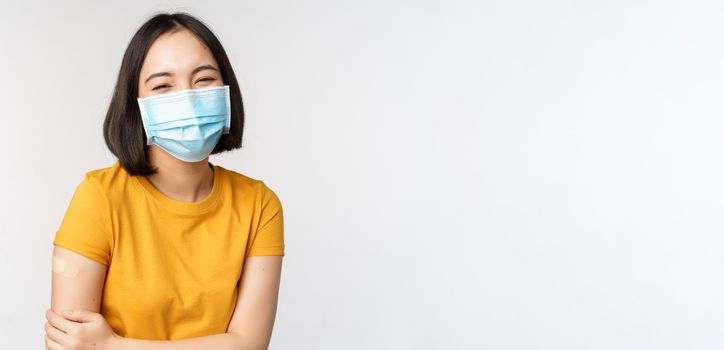 Healthy and happy asian woman in medical face mask, has band aid on shoulder after covid-19 vaccination, got vaccinated from omicron delta variant, whtie background.