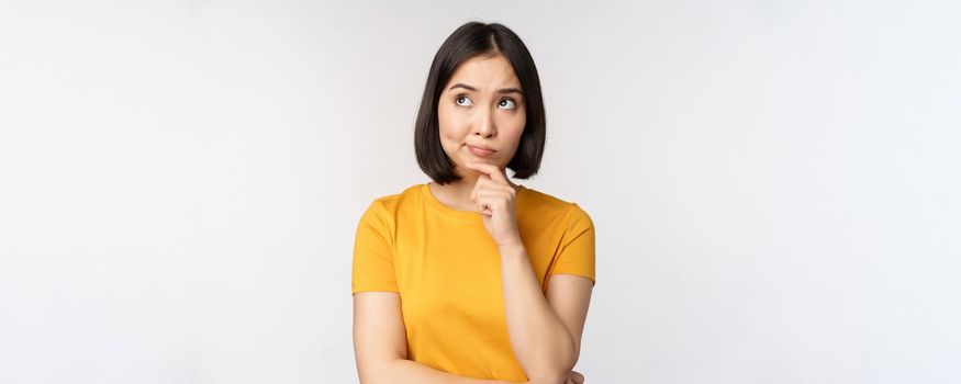 Image of thinking asian woman, looking aside and pondering, making decision, standing in yellow tshirt over white background.
