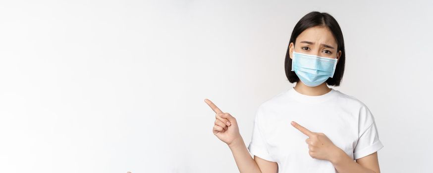 Sad asian woman in medical mask, pointing fingers left, frowning and looking upset, complaining, demonstrating banner, standing over white background.