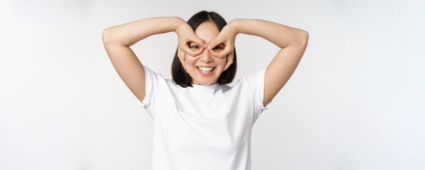 Funny young asian woman, korean girl making eyes glasses gesture, looking happy at camera, standing over white background.