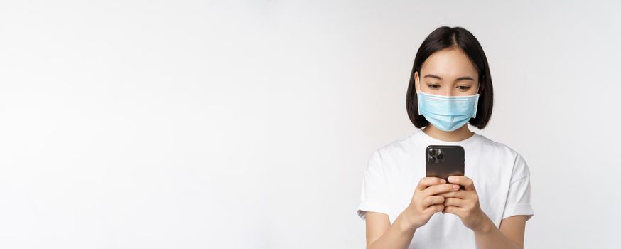 Health, covid and mobile concept. Young asian woman in medical face mask, looking at smartphone screen, using phone app, shopping online, white background.