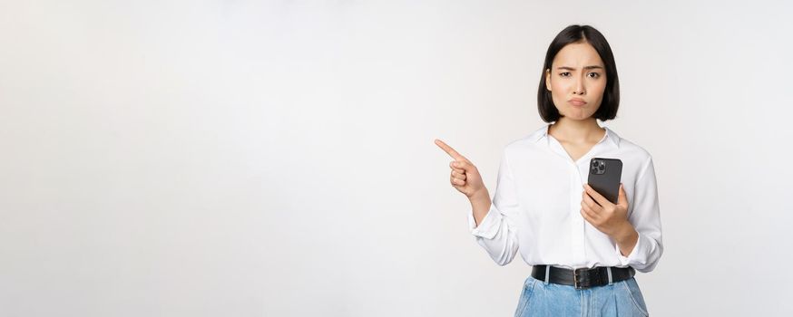 Skeptical asian modern girl, holding smartphone, pointing finger left at empty space and frowing hesitant, has doubts, standing over white background.