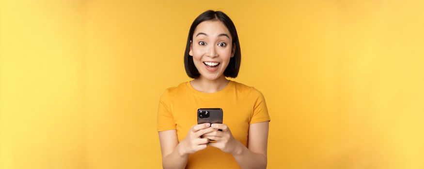 Happy asian girl smiling, standing with black mobile phone, standing against yellow background. Copy space