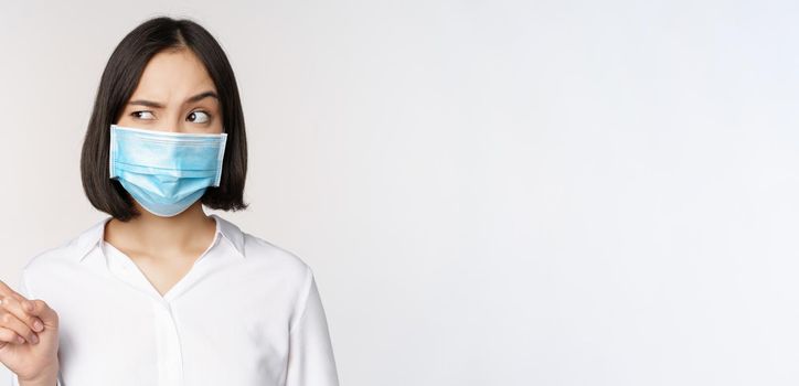 Image of young asian woman in medical face mask, pointing left and looking with suspicious confused expression, standing over white background.