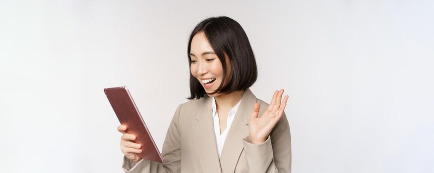Portrait of smiling asian businesswoman video chat on digital tablet, waving hand at gadget screen, standing in suit over white background.