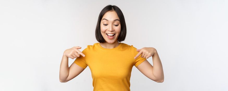 Happy asian girl pointing fingers down, looking at announcement, banner or advertisment, standing in yellow tshirt over white background.