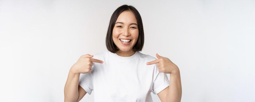 Happy and confident asian woman, student smiling and pointing at herself, self-promoting, showing logo on t-shirt, standing over white background.