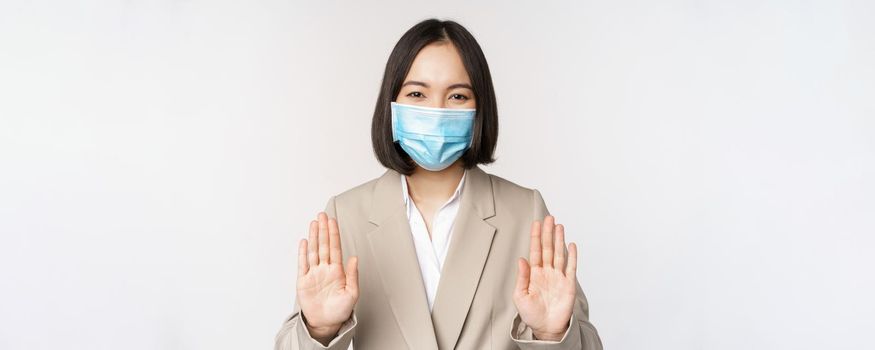 Coronavirus and work concept. Portrait of asian female office lady, woman at workplace wearing medical face mask and showing stop, prohibition gesture, white background.